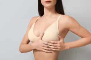 Young woman examining her breast against a grey background, symbolizing evaluation before or after fat transfer breast augmentation treatment.