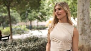 Thoughtful woman in sleeveless top standing in a sunny park, representing confidence and healing following male-to-female top surgery.