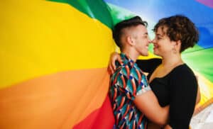 LGBTQ couple embracing affectionately against rainbow pride flag, smiling and bonding with love and support.